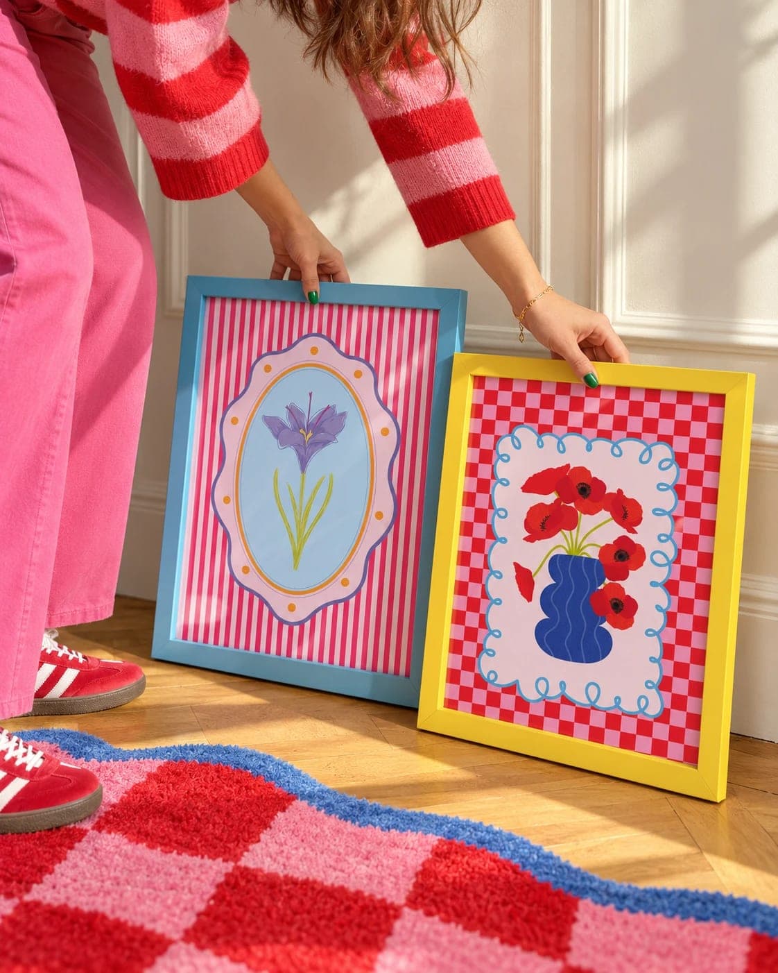 Iris and Dahnon prints in blue and yellow frames being placed against a wall, above a red and pink checkered rug